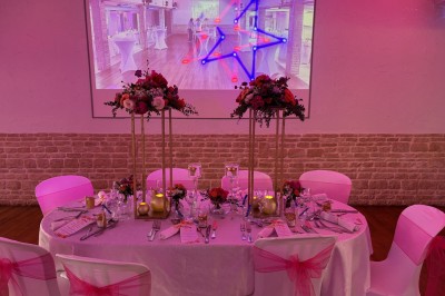 Salle de réception décorée avec des tables rondes dressées, éclairage rose et chaises blanches.
