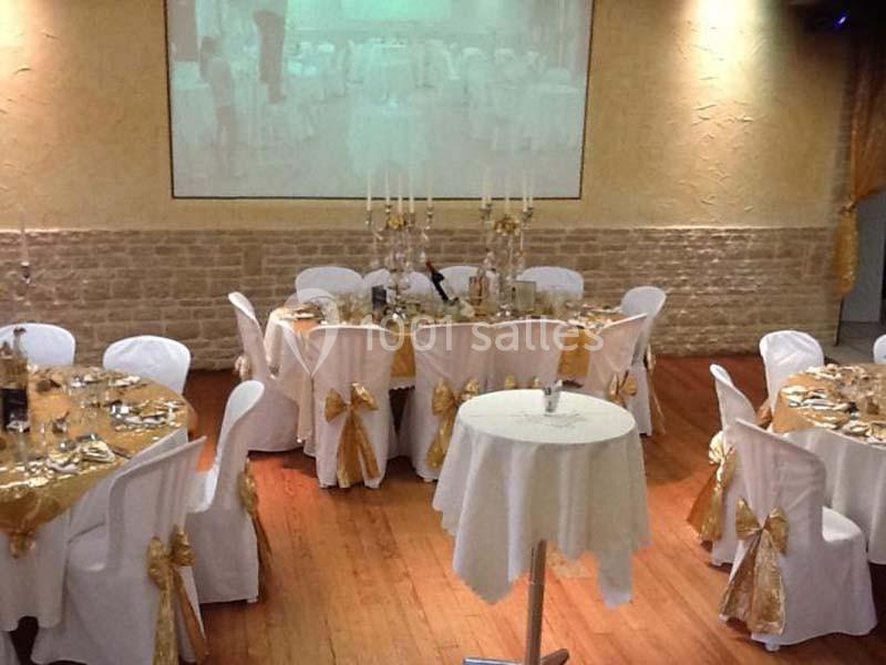Salle de réception décorée avec des tables rondes, nappes blanches et dorées, chandeliers et chaises ornées de nœuds.