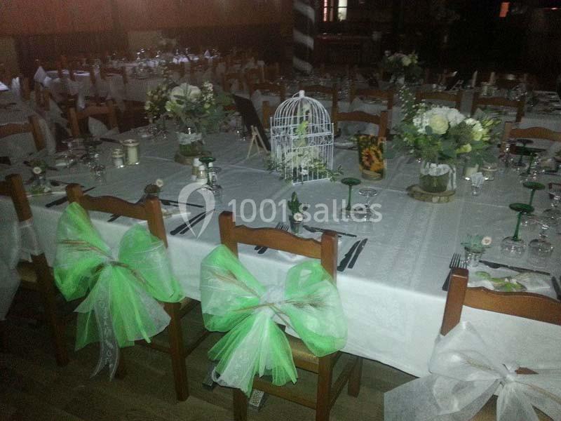 Salle décorée pour un événement avec des tables dressées, des chaises ornées de nœuds verts et une cage blanche en centre de…