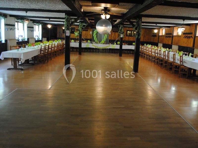 Salle de réception avec tables alignées et dressées, décor rustique en bois et éclairage suspendu.