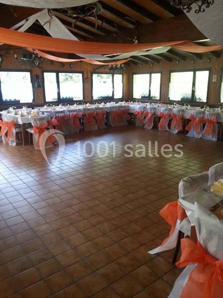 Salle décorée pour un événement avec des tables alignées, nappes blanches et chaises ornées de rubans orange.
