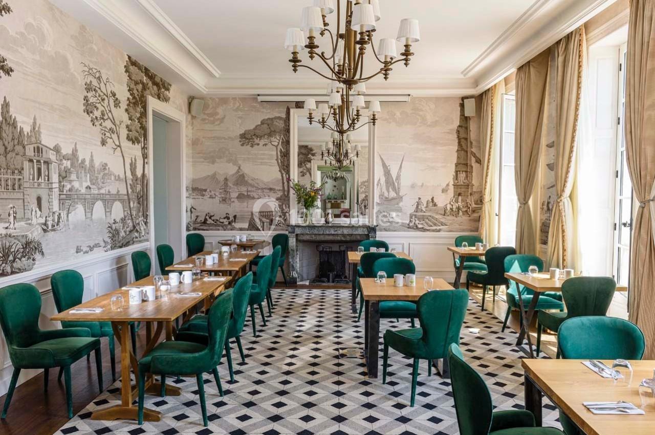 Salle de restaurant lumineuse avec des chaises vertes, tables en bois, fresques murales et un lustre central.
