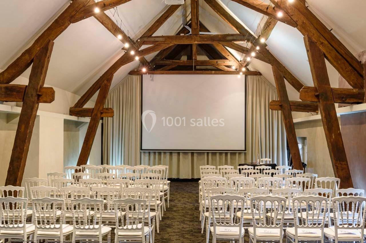 Salle de conférence avec poutres apparentes, écran de projection, rideaux beiges et chaises blanches alignées.