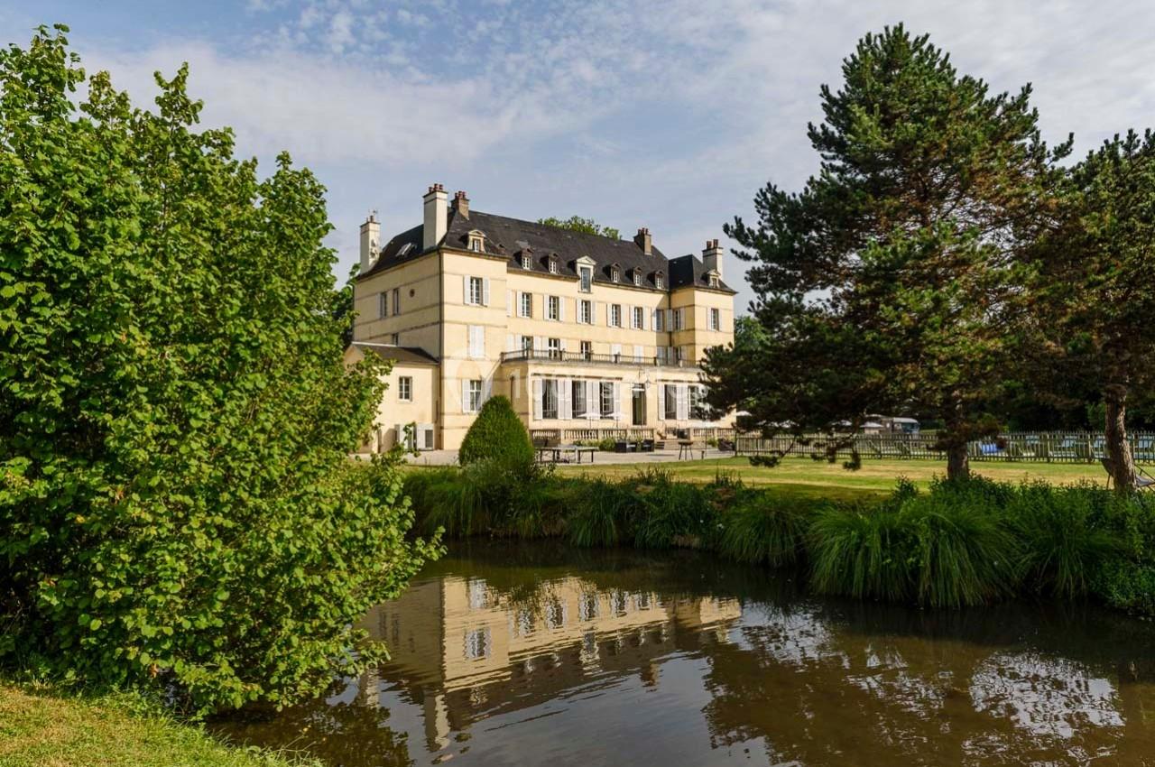 Façade d'un grand bâtiment historique entouré d'arbres, avec un étang reflétant son image au premier plan.