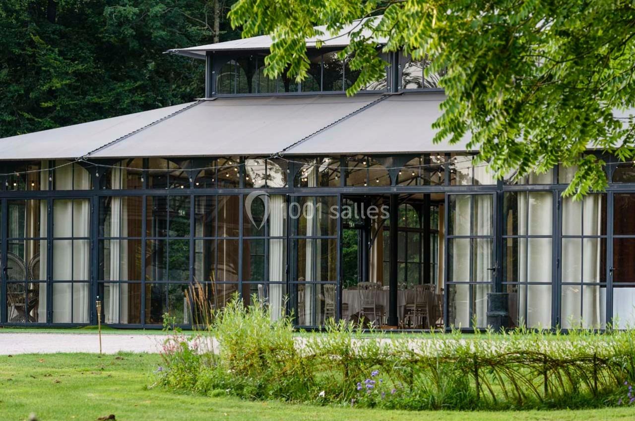 Vue extérieure d'une verrière moderne entourée de verdure, avec des tables dressées visibles à l'intérieur.