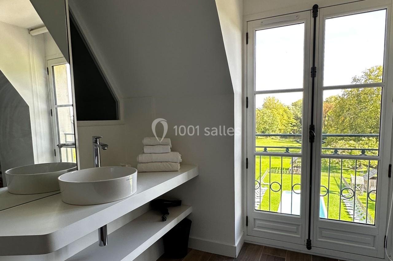 Salle de bain lumineuse avec vasque blanche, serviettes pliées sur une étagère et vue sur un jardin par une porte-fenêtre.