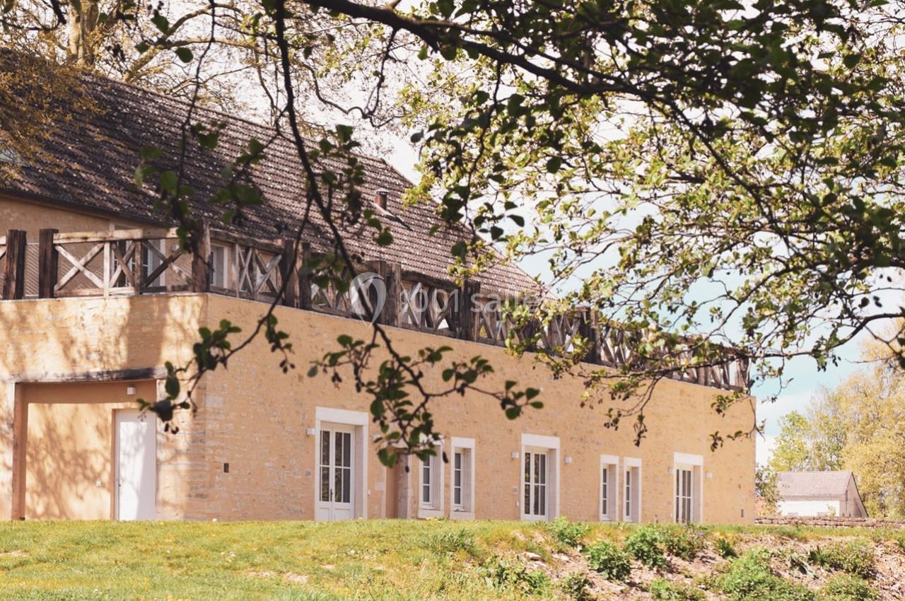 Bâtiment en pierre beige avec fenêtres blanches, entouré de verdure et partiellement ombragé par des branches d'arbre.