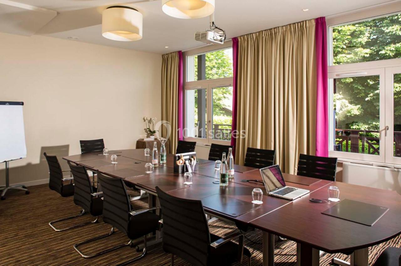Salle de réunion lumineuse avec une grande table, des chaises noires, un tableau blanc et des fenêtres donnant sur un espace…