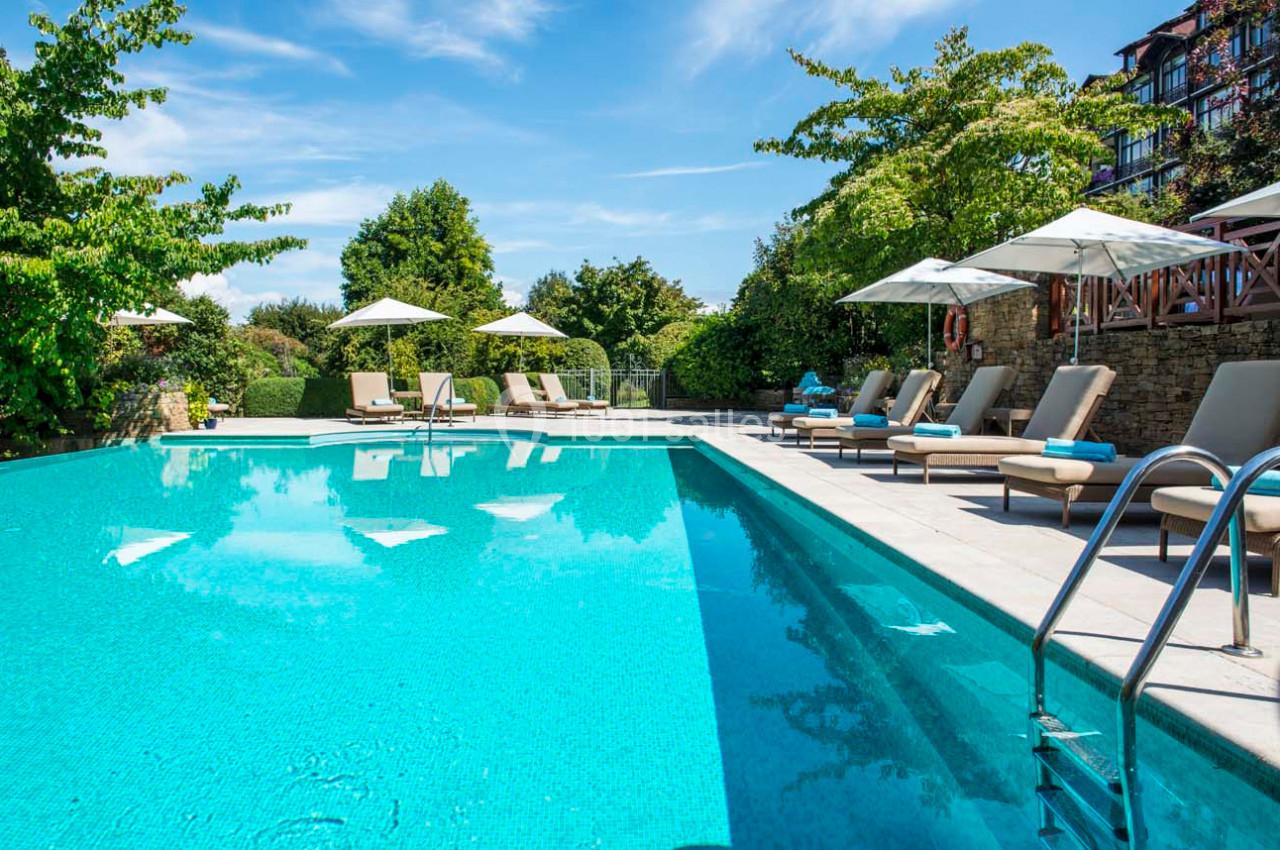 Piscine extérieure entourée de transats, parasols et végétation sous un ciel bleu clair.
