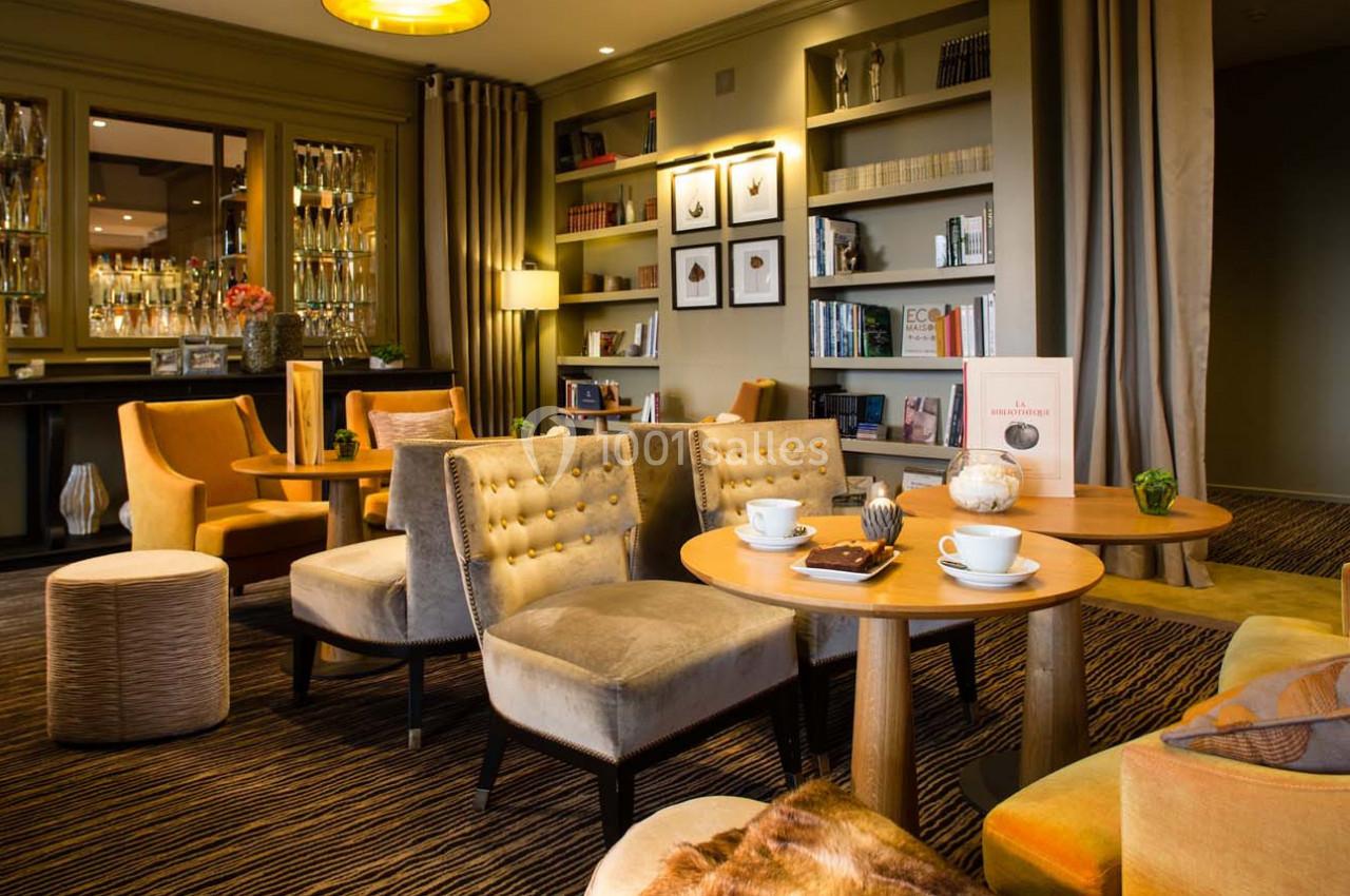 Salon élégant avec fauteuils rembourrés, tables en bois, bibliothèque et éclairage chaleureux.