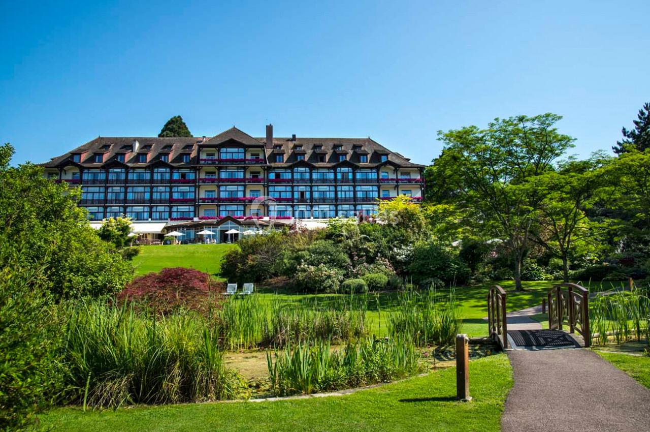Hôtel à colombages entouré de jardins verdoyants avec un petit pont en bois traversant un espace paysager.