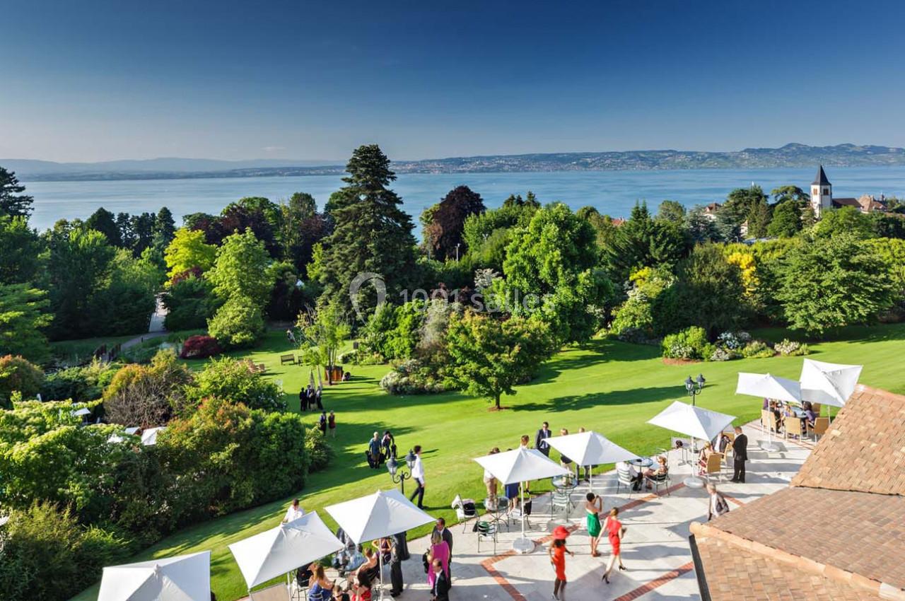 Jardin verdoyant avec vue sur un lac, des montagnes à l'horizon et des parasols blancs sur une terrasse animée.