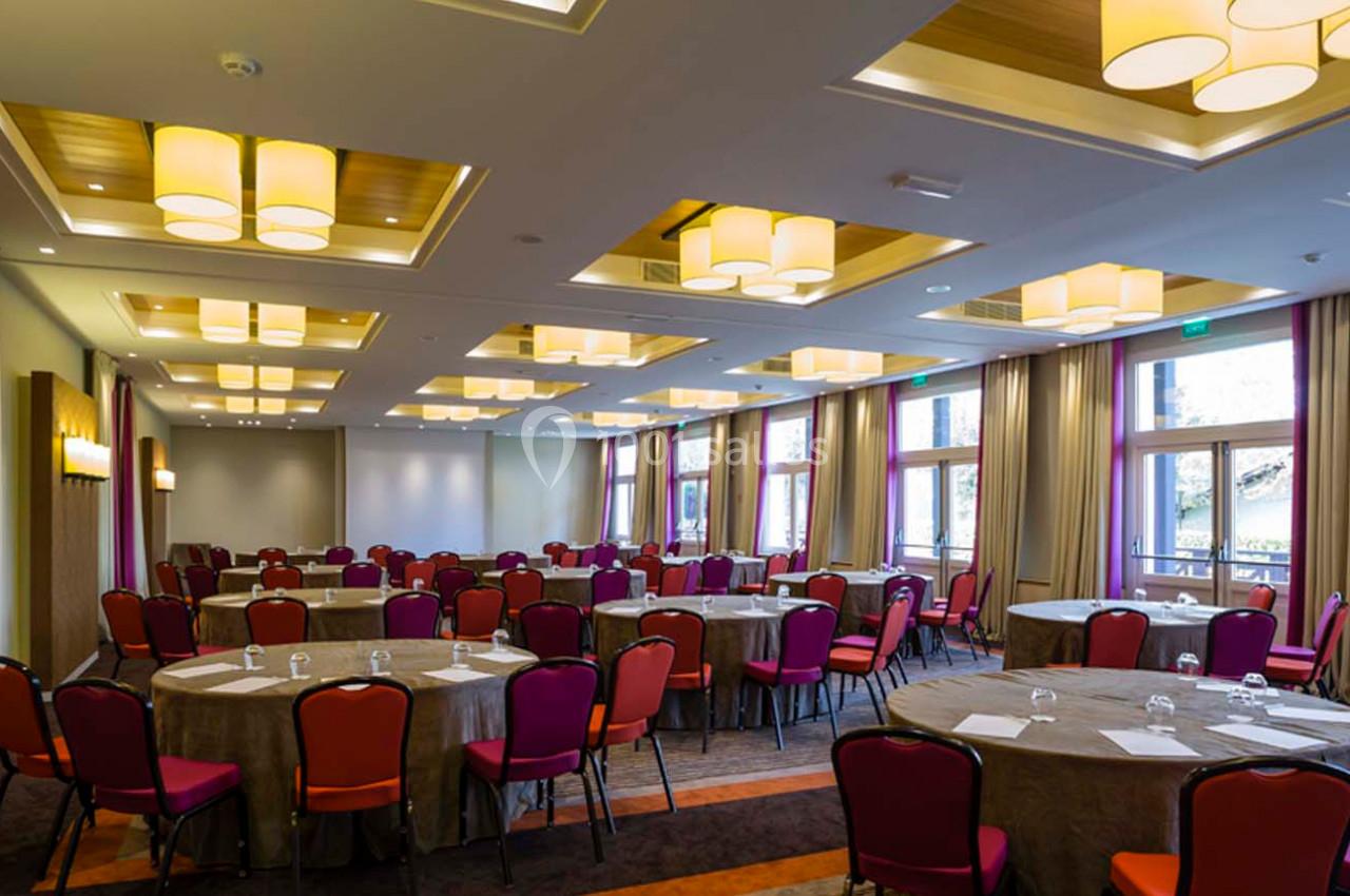 Salle de conférence lumineuse avec des tables rondes dressées, des chaises rouges et de grandes fenêtres.