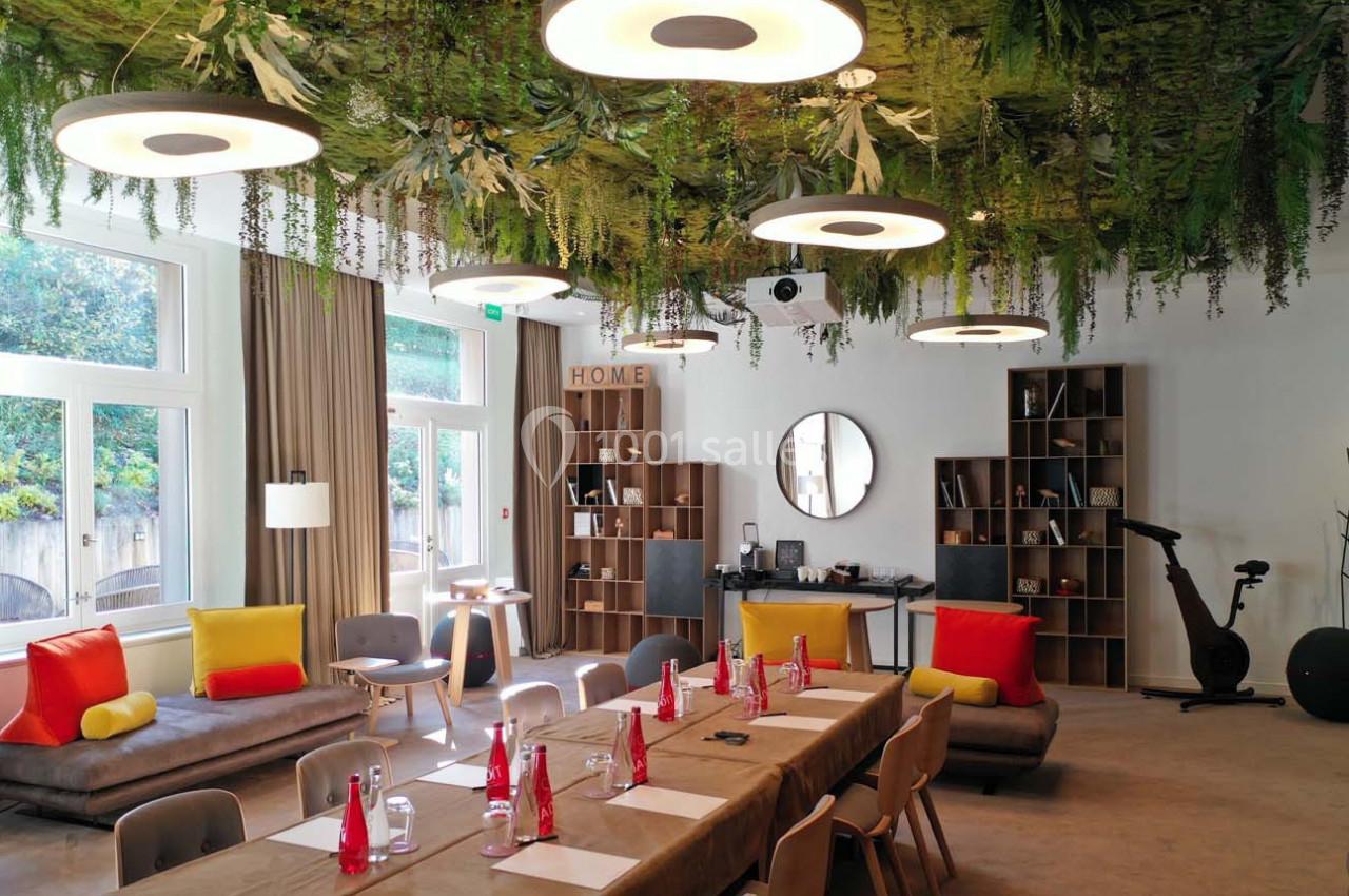 Salle lumineuse avec une grande table dressée, plafond végétalisé, canapés colorés et équipements de détente.