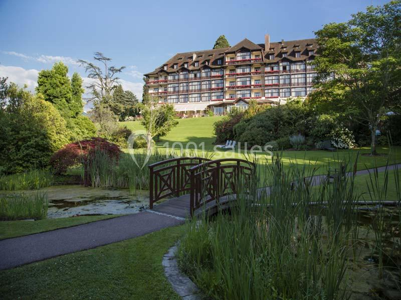 Jardin verdoyant avec un petit pont en bois, étang et grand bâtiment à plusieurs étages en arrière-plan.