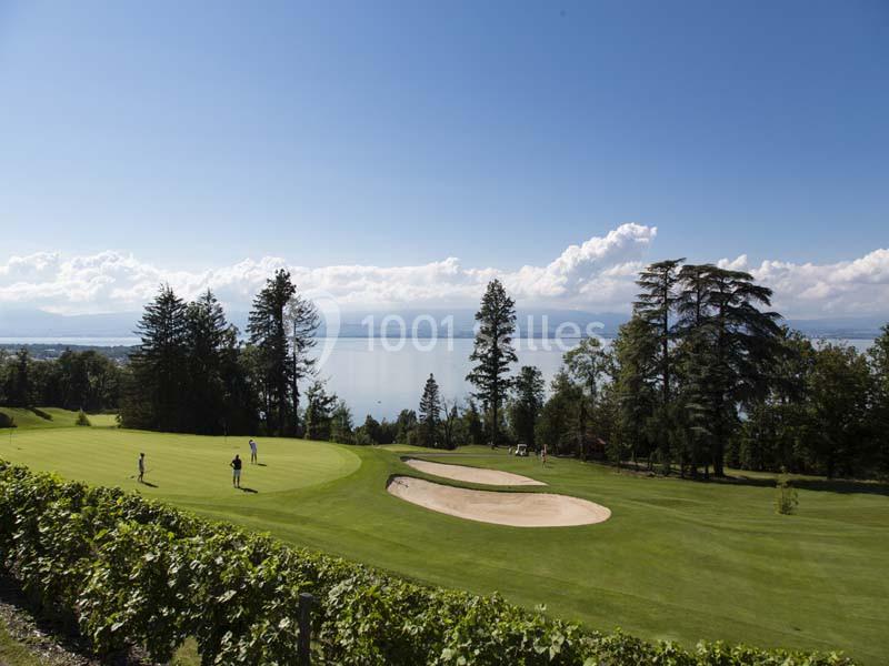 Vue d'un terrain de golf verdoyant avec des bunkers, entouré de vignes et surplombant un lac sous un ciel dégagé.