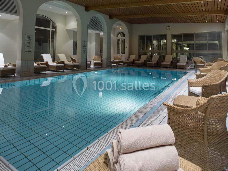 Piscine intérieure avec carrelage bleu, entourée de chaises longues et de fauteuils en osier sous un plafond en bois.