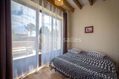 Chambre lumineuse avec lit double, rideaux transparents, plafond en poutres apparentes et vue sur une terrasse ensoleillée.