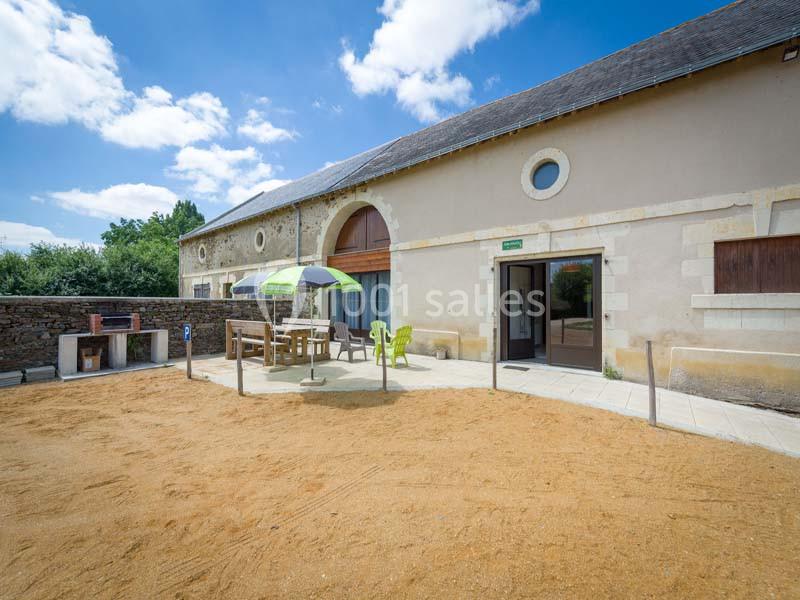 Cour extérieure ensoleillée avec une table, des chaises sous un parasol, et un bâtiment en pierre en arrière-plan.