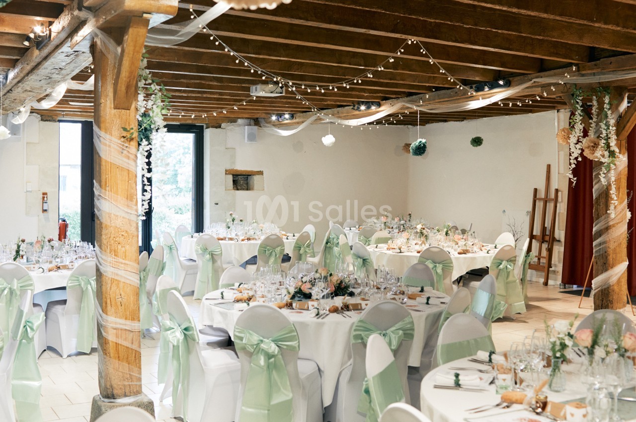 Salle de réception décorée pour un mariage, avec tables rondes, nappes blanches, chaises ornées de rubans verts et…