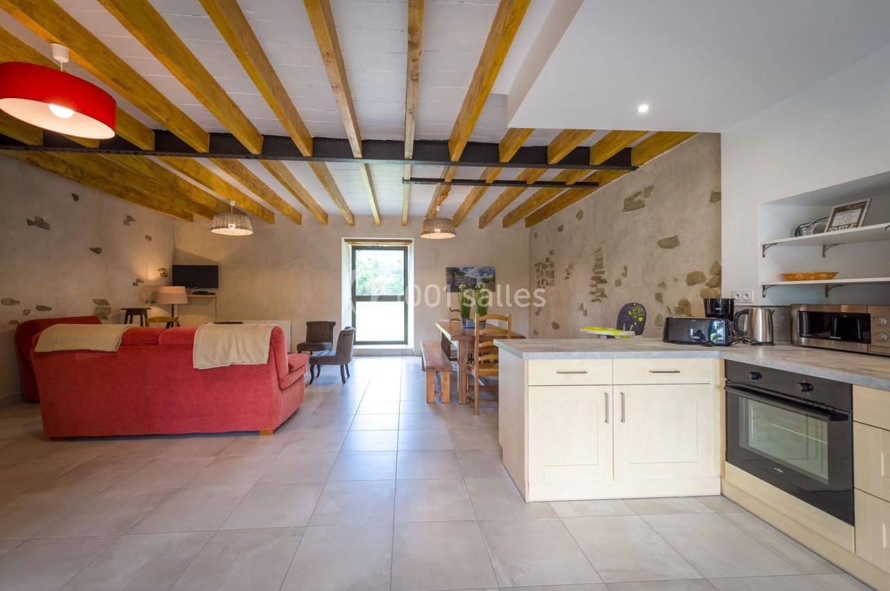 Pièce de vie lumineuse avec cuisine équipée, table à manger, canapé rouge et poutres apparentes au plafond.