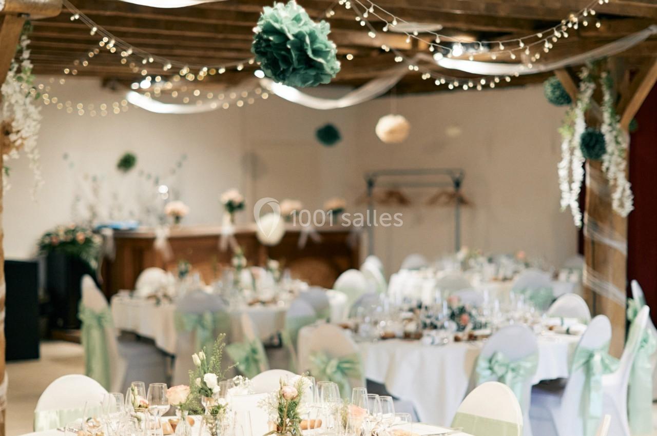 Salle de réception décorée pour un mariage, avec tables rondes, chaises ornées de rubans verts et guirlandes lumineuses.