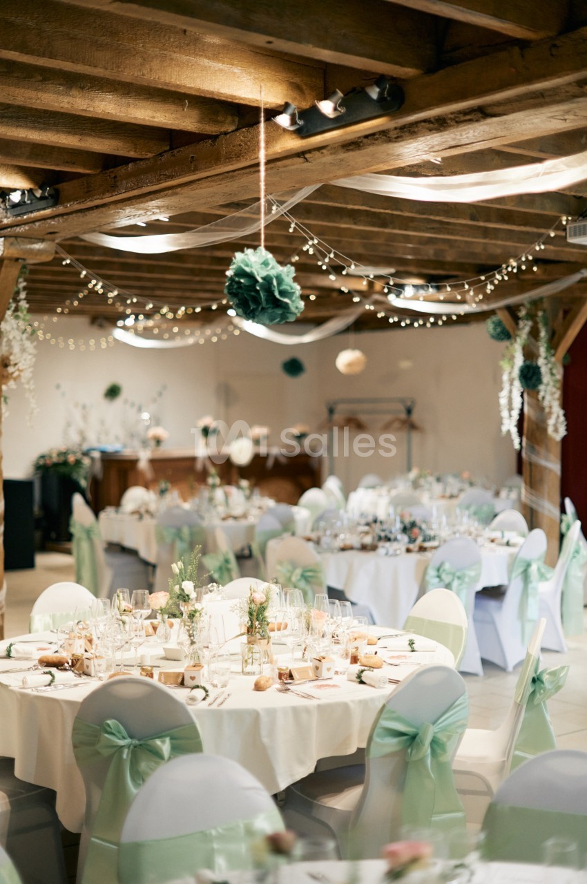 Salle de réception décorée pour un mariage, avec tables rondes, chaises ornées de rubans verts et guirlandes lumineuses.