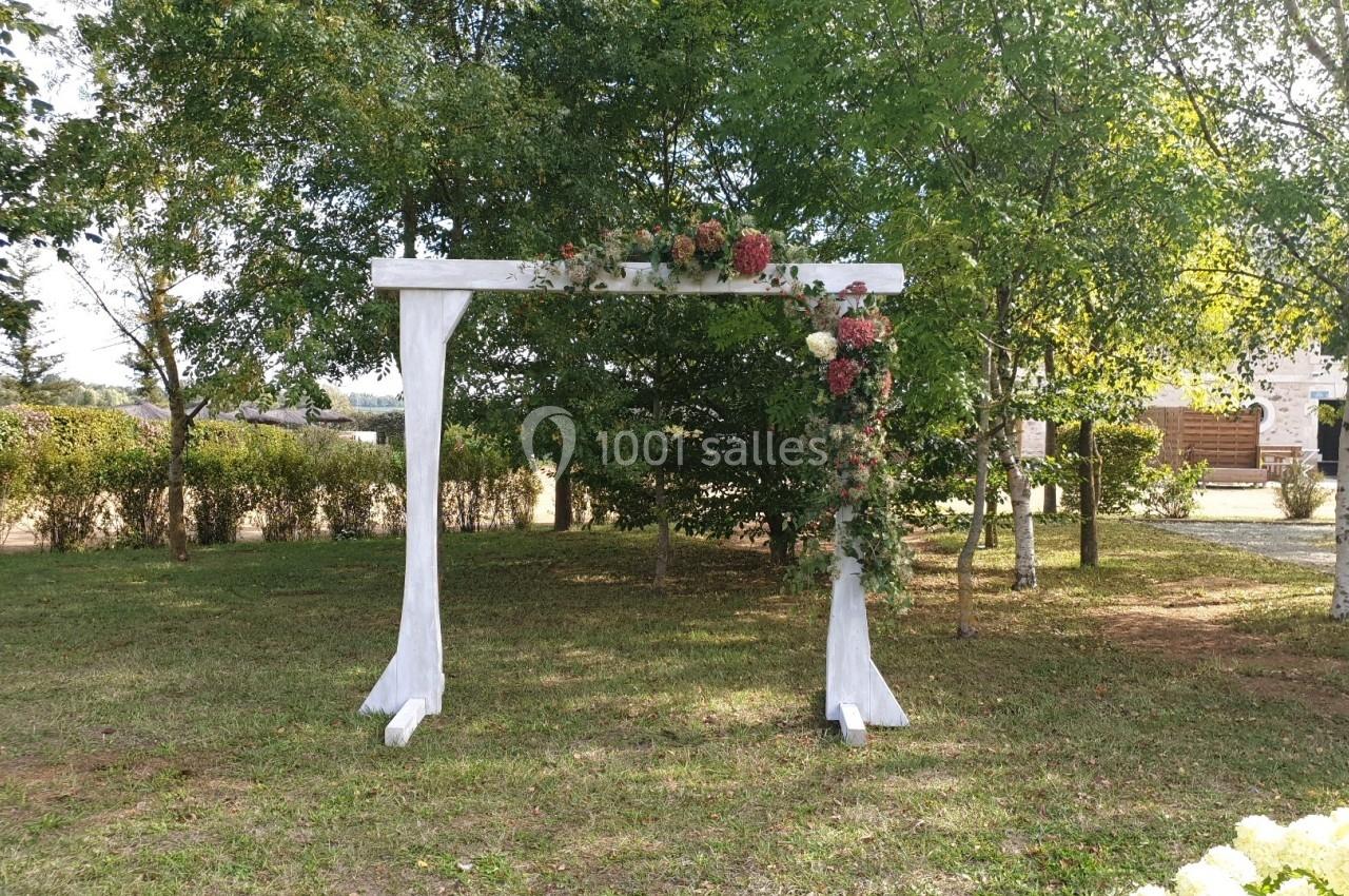 Arche en bois blanc décorée de fleurs rouges et roses, installée dans un jardin verdoyant entouré d'arbres.
