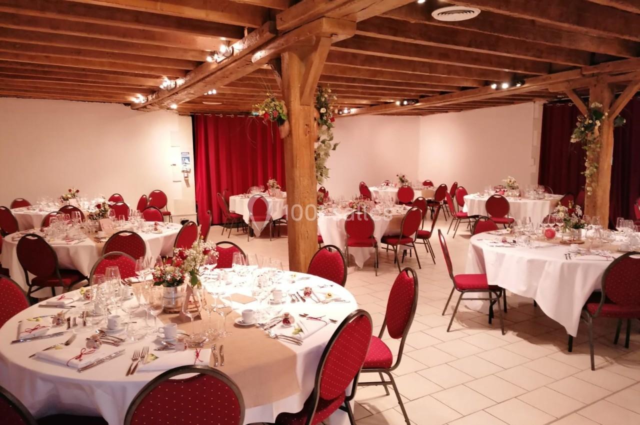 Salle de réception avec tables rondes dressées, nappes blanches, chaises rouges et décoration florale.