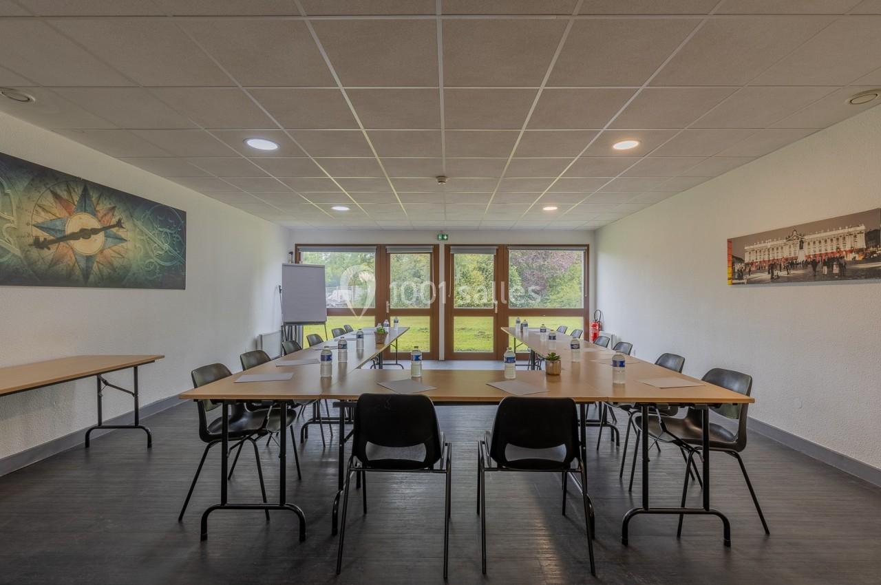 Salle de réunion lumineuse avec tables en U, chaises noires, bouteilles d'eau et vue sur un espace vert à travers des baies…
