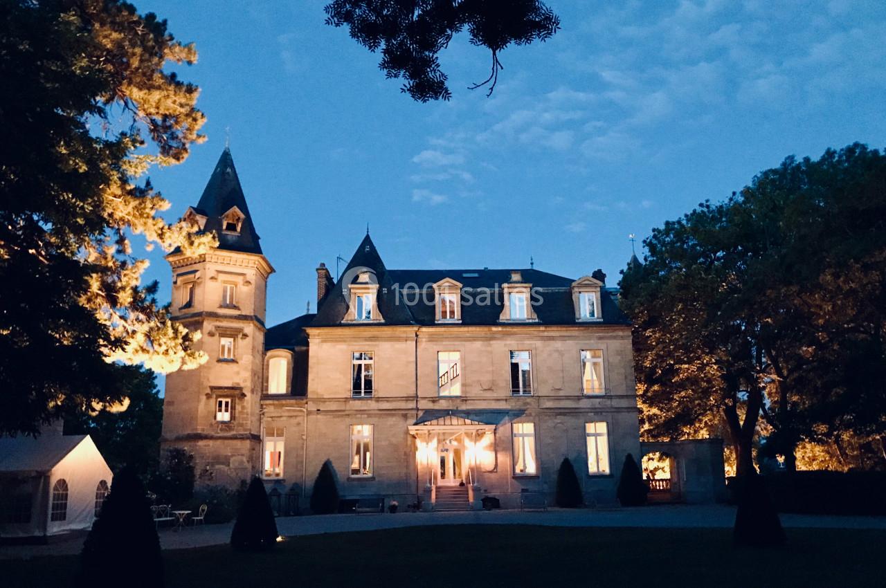 Façade éclairée d'un château en soirée, entouré d'arbres et d'un ciel légèrement nuageux.