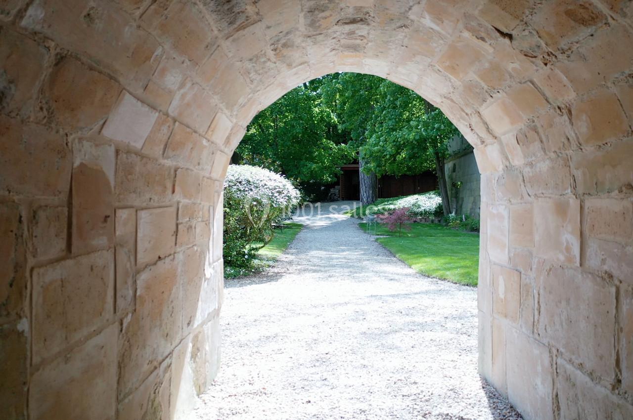 Vue depuis une arche en pierre donnant sur un chemin gravillonné entouré de verdure et de fleurs.