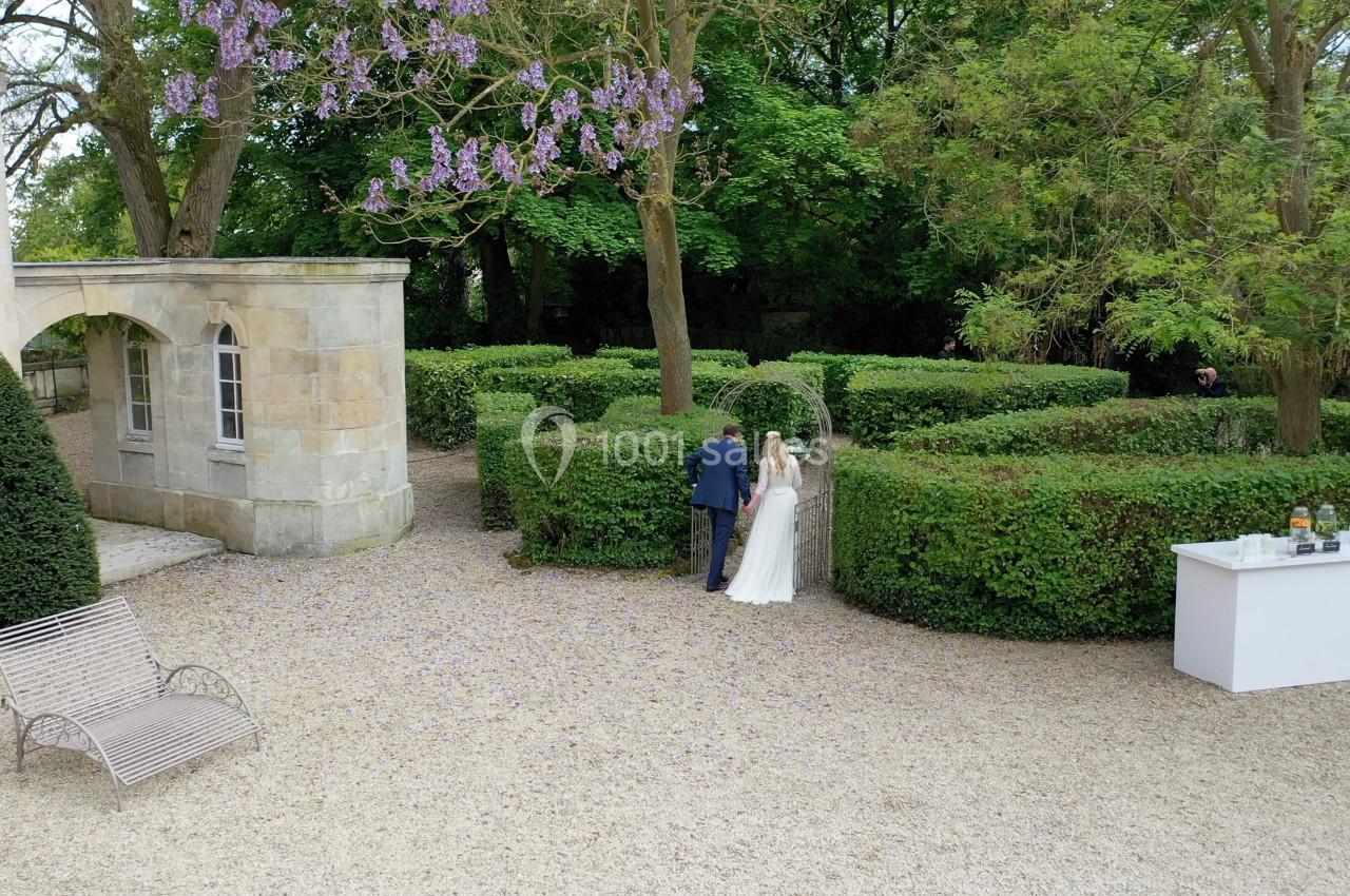 Un couple en tenue de mariage se tient près d'une haie dans un jardin arboré, avec une table blanche à proximité.