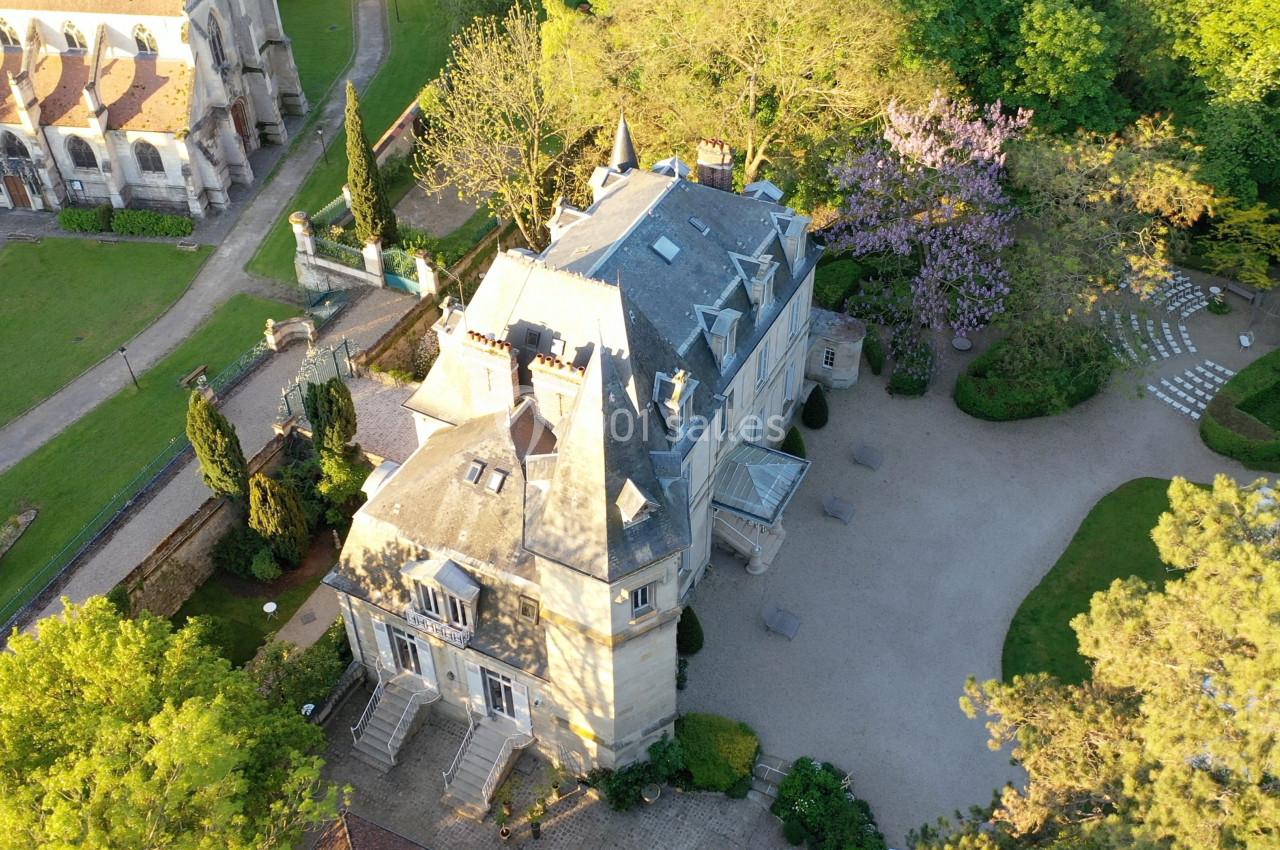 Vue aérienne d'un manoir entouré de jardins verdoyants, avec une église visible en arrière-plan à gauche.