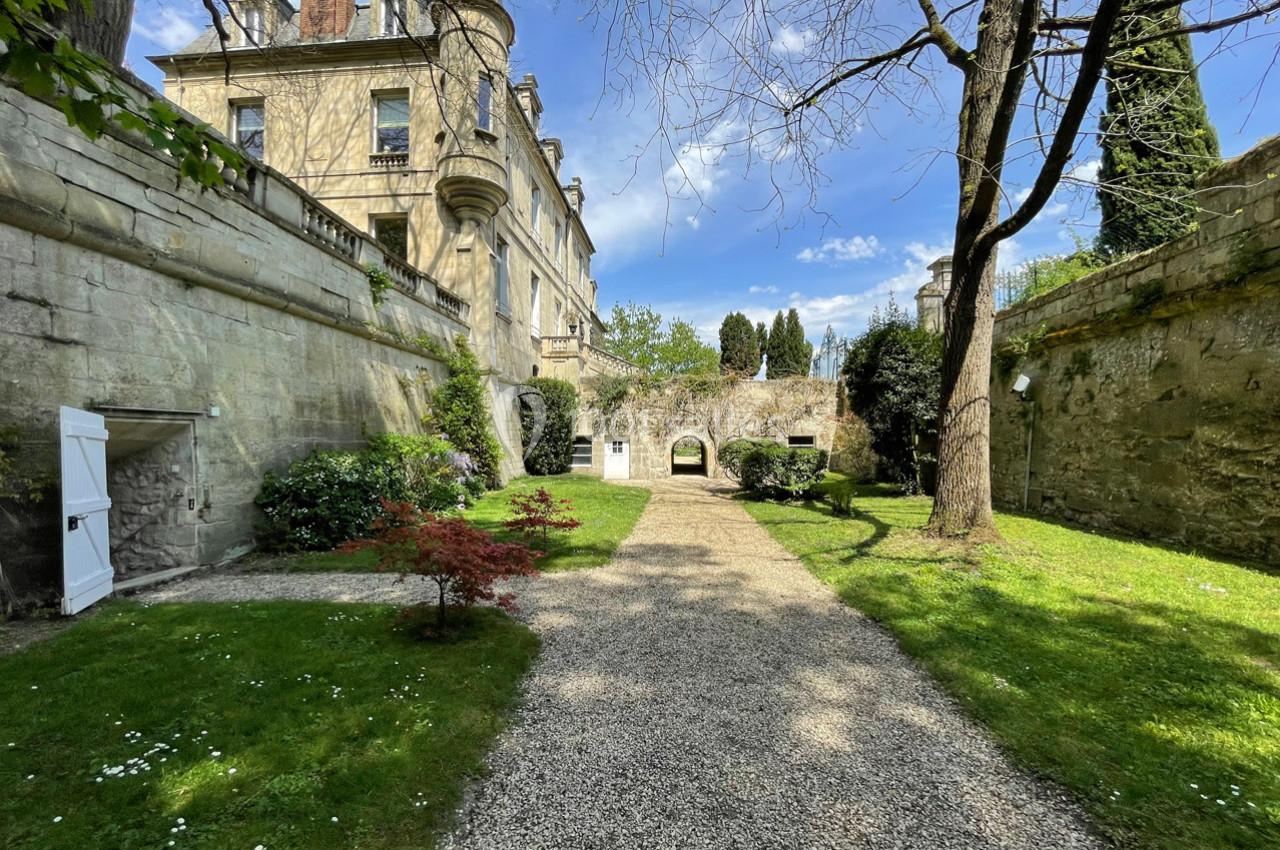 Jardin avec allée gravillonnée, arbres, pelouse et murs en pierre entourant un bâtiment ancien sous un ciel bleu.