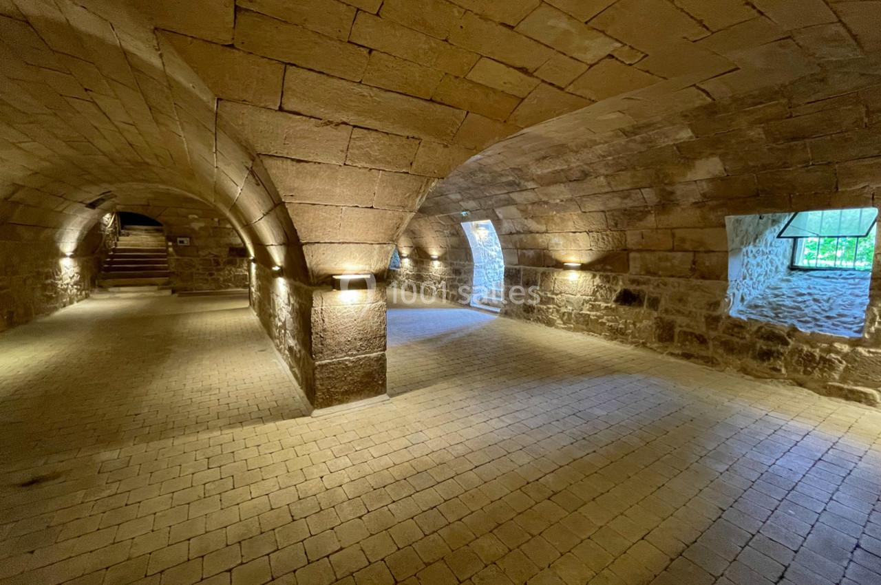 Voûtes en pierre éclairées par des lumières murales, avec des fenêtres laissant entrer la lumière naturelle.