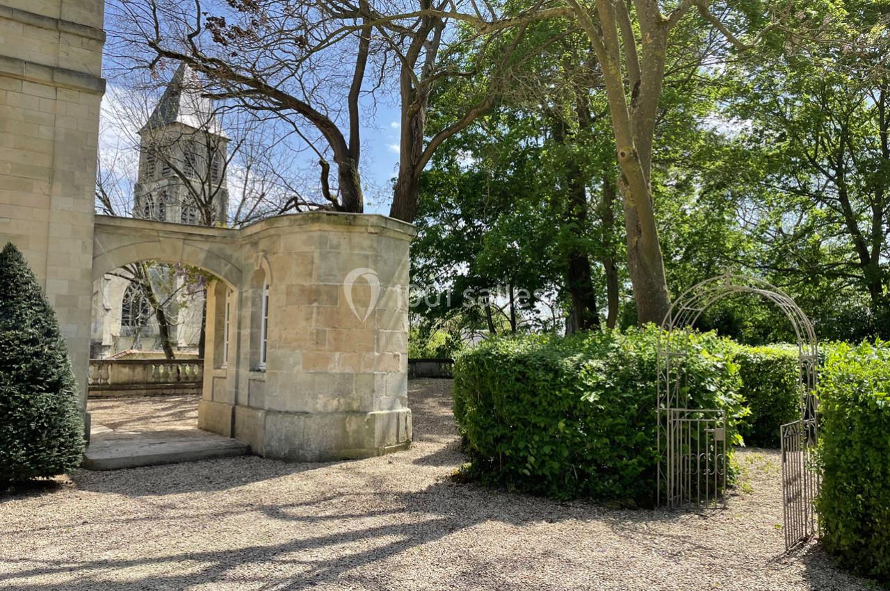 Allée gravillonnée bordée de haies et d'arbres, avec une arche métallique et un bâtiment en pierre à gauche.
