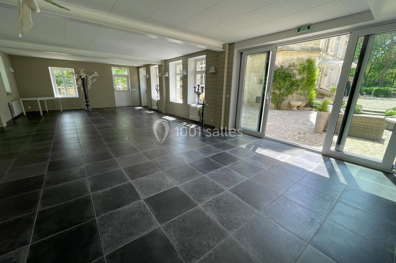 Salle lumineuse avec carrelage noir, grandes fenêtres et portes vitrées donnant sur une cour extérieure.