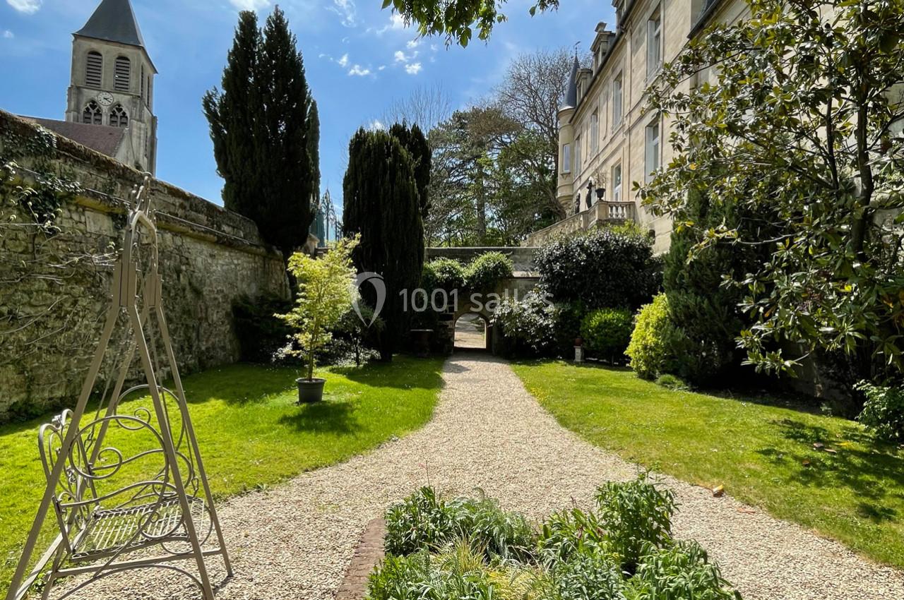 Jardin avec allée gravillonnée, végétation variée, bancs, et clocher d'église visible en arrière-plan.