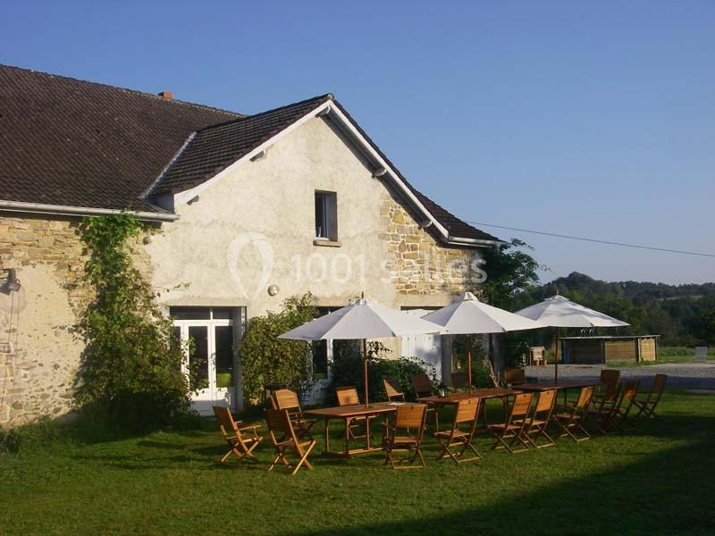 Maison en pierre avec toit en pente, terrasse aménagée avec tables et chaises en bois sous des parasols blancs.