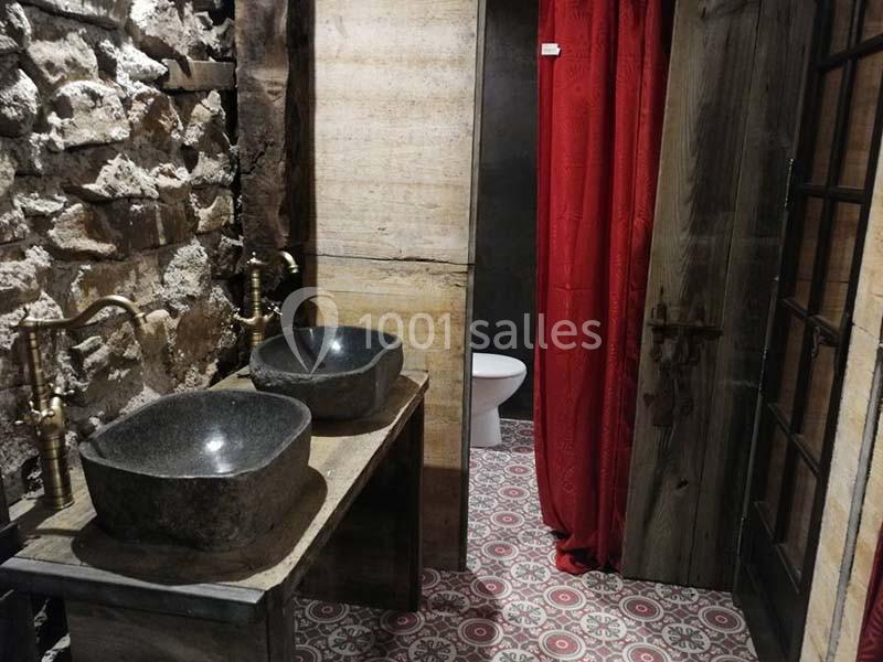 Salle de bain rustique avec murs en pierre, vasques en pierre noire, rideau rouge et sol à motifs géométriques.