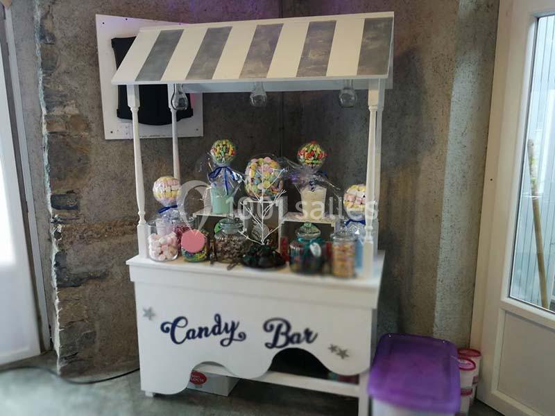 Chariot blanc décoré avec des bonbonnières remplies de friandises colorées, installé devant un mur en pierre.