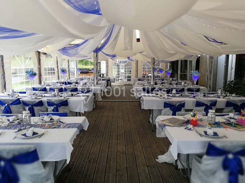 Salle de réception décorée de nappes blanches et de rubans bleus, avec des tables dressées sous une tente.