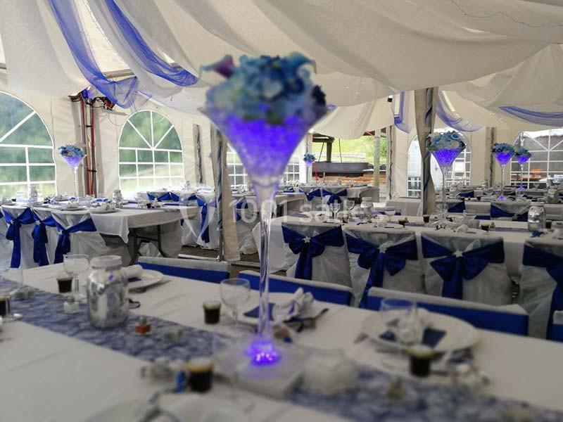 Salle de réception décorée avec des nappes blanches, des rubans bleus et des centres de table lumineux.