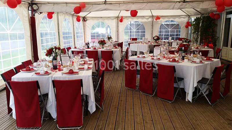 Salle décorée pour un événement avec des tables dressées, nappes blanches, chaises rouges et ballons rouges suspendus.