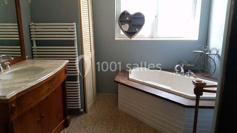 Salle de bain avec baignoire d'angle, meuble en bois avec lavabo, miroir, radiateur et fenêtre avec décoration en forme de…