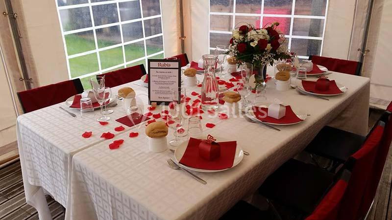 Table dressée avec nappes blanches, pétales rouges, verres, assiettes, serviettes pliées et un bouquet de fleurs au centre.
