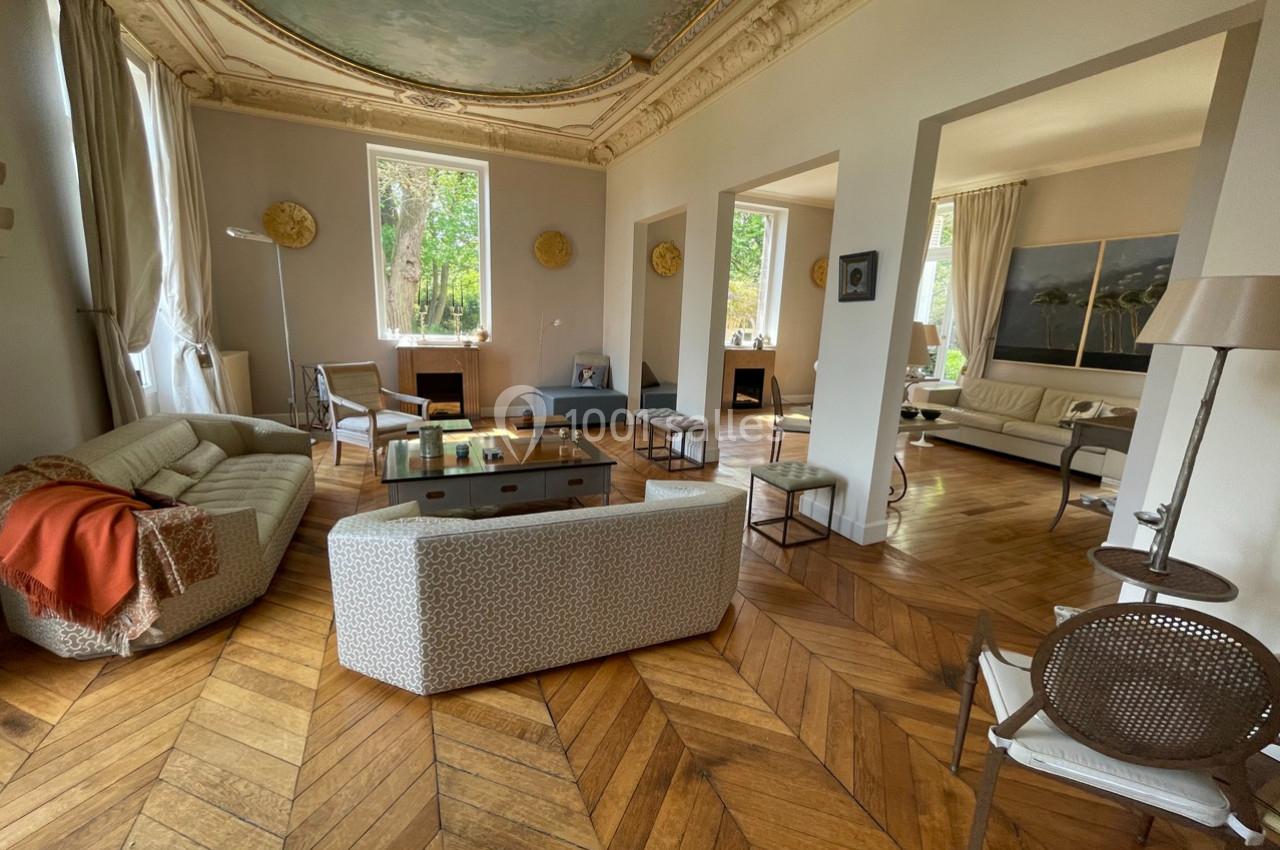 Salon lumineux avec parquet en chevrons, mobilier moderne, hauts plafonds ornés et grandes fenêtres donnant sur un jardin.