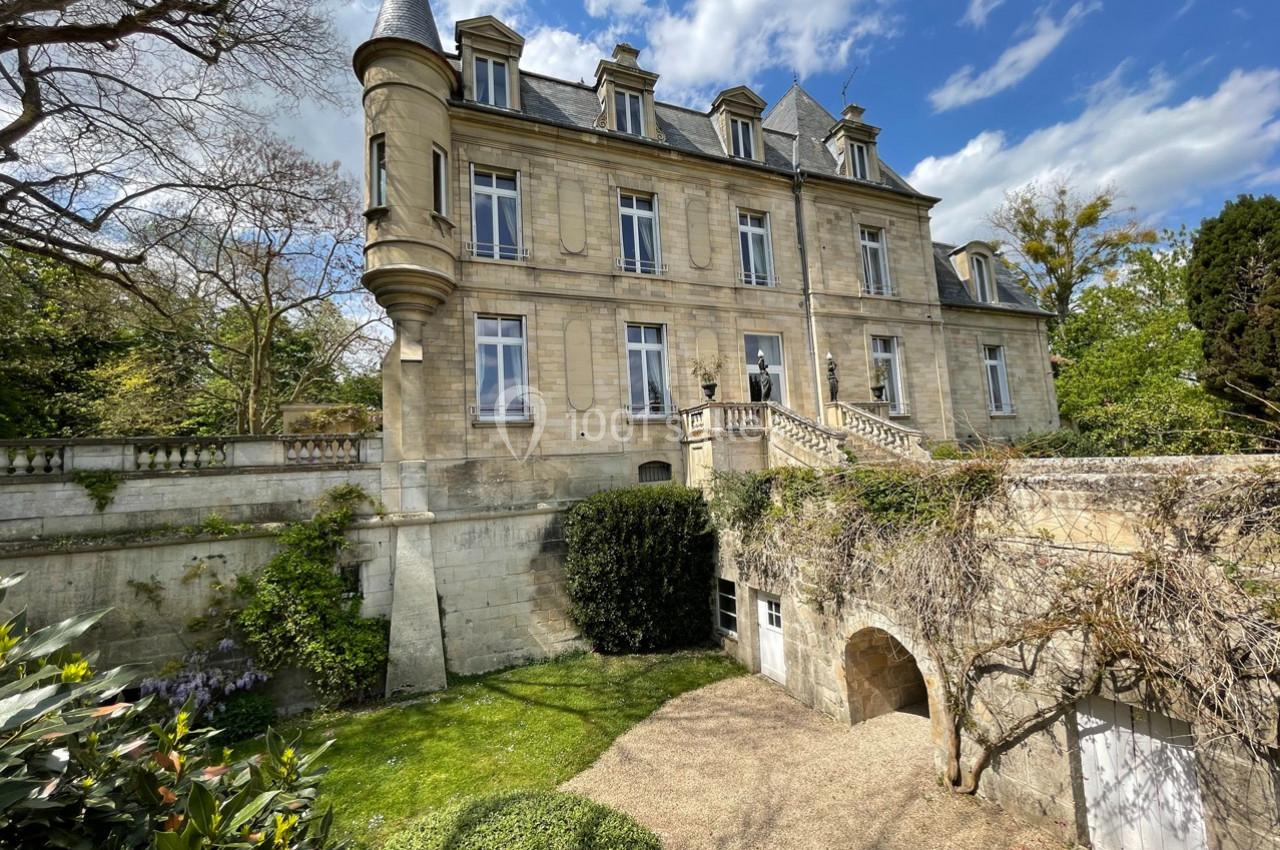 Façade d'un château en pierre avec tourelle, entouré de verdure et d'un jardin sous un ciel partiellement nuageux.