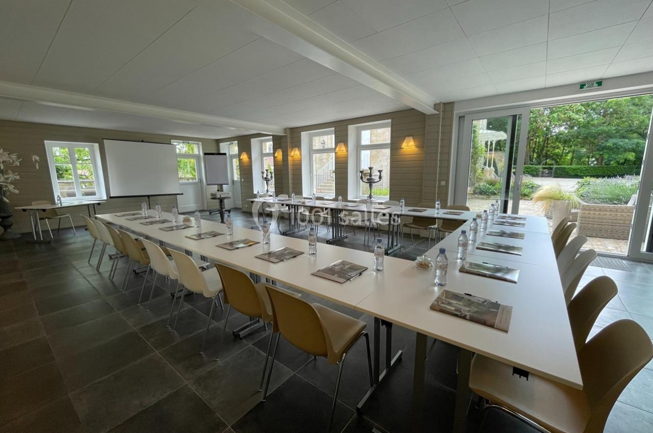 Salle de réunion lumineuse avec tables disposées en U, chaises, écran de projection et vue sur un jardin extérieur.